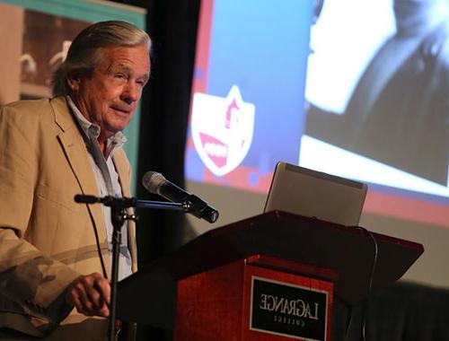 Professor John Lawrence stands behind a podium on stage as he speaks about his 50 years of service a the college